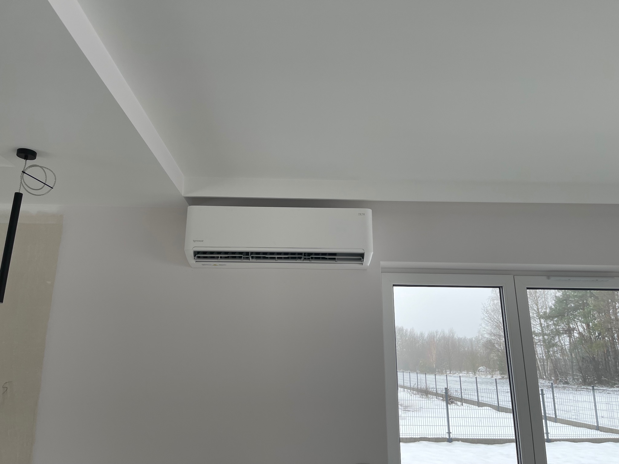 Biały klimatyzator Rotenso Neo, zamontowany na ścianie w jasnym pomieszczeniu z widokiem na zimowy krajobraz za oknem. Minimalistyczny design, nowoczesne wykończenie.