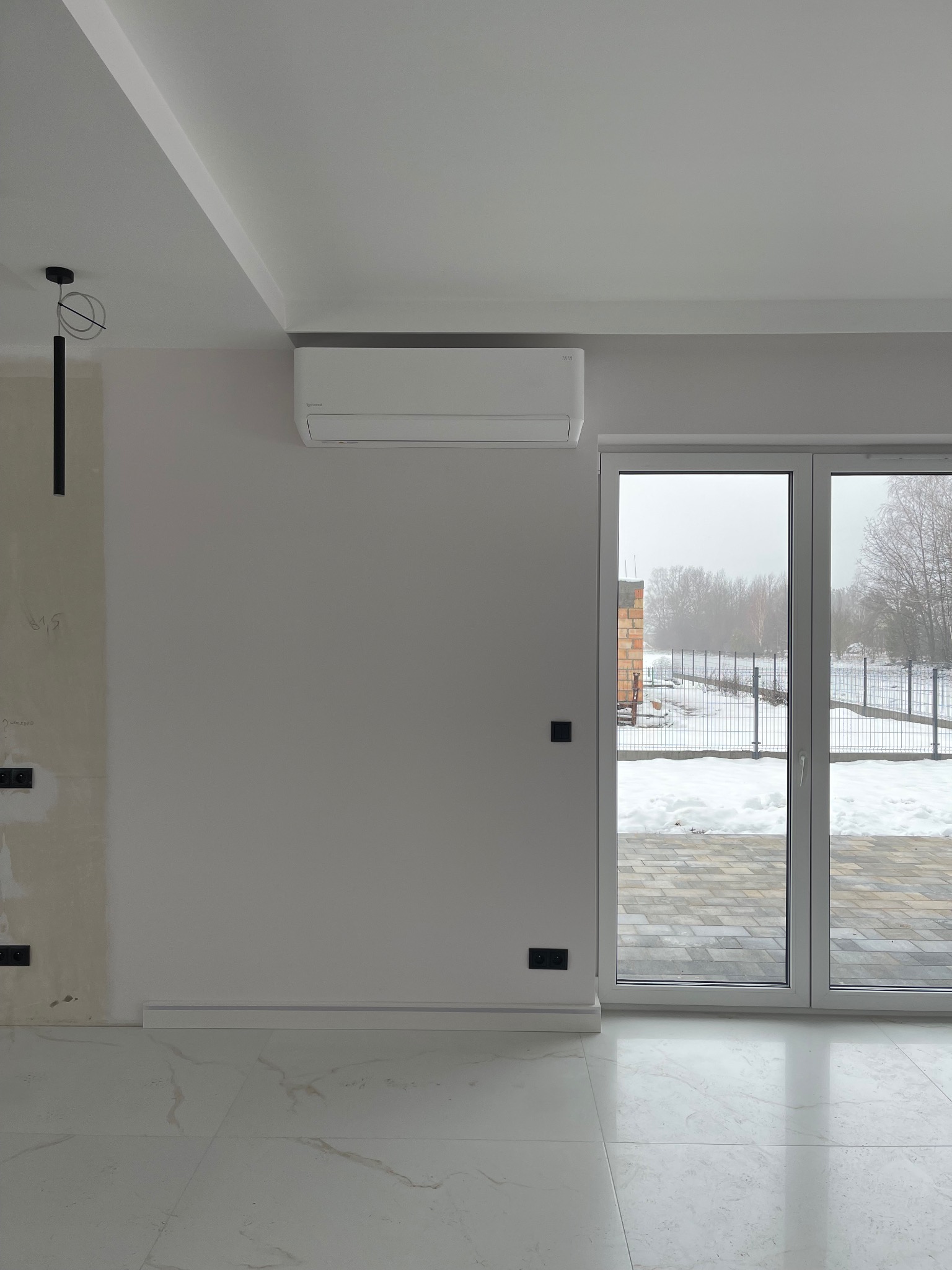 Biały klimatyzator na ścianie w jasnym wnętrzu, widoczne okno z widokiem na zaśnieżony ogród. Minimalistyczny design, nowoczesne wykończenie, widoczne gniazdka elektryczne.