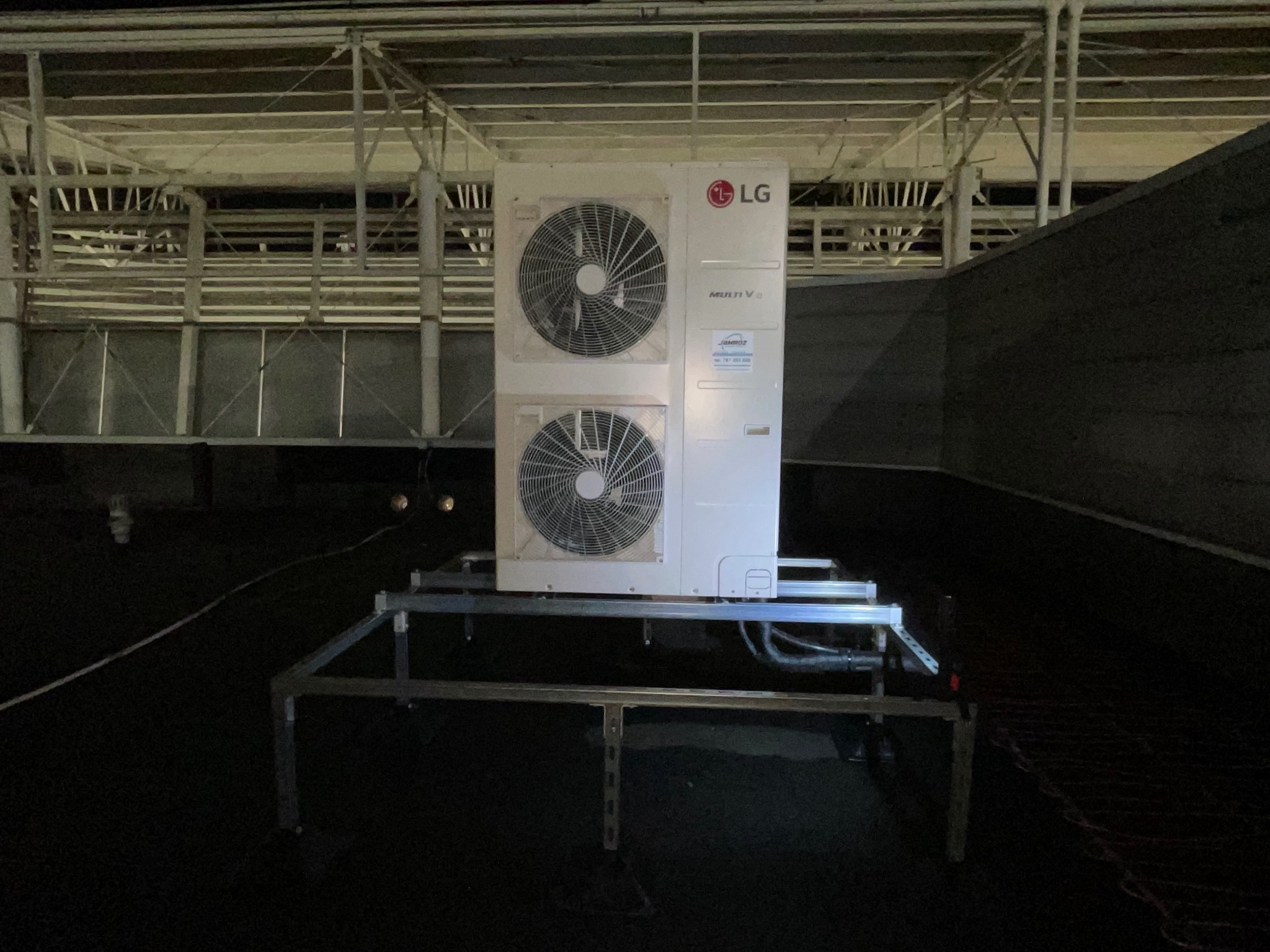 Biała jednostka zewnętrzna klimatyzacji LG Multi V, zamontowana na metalowej konstrukcji na dachu budynku, widoczna pod światłem reflektorów.