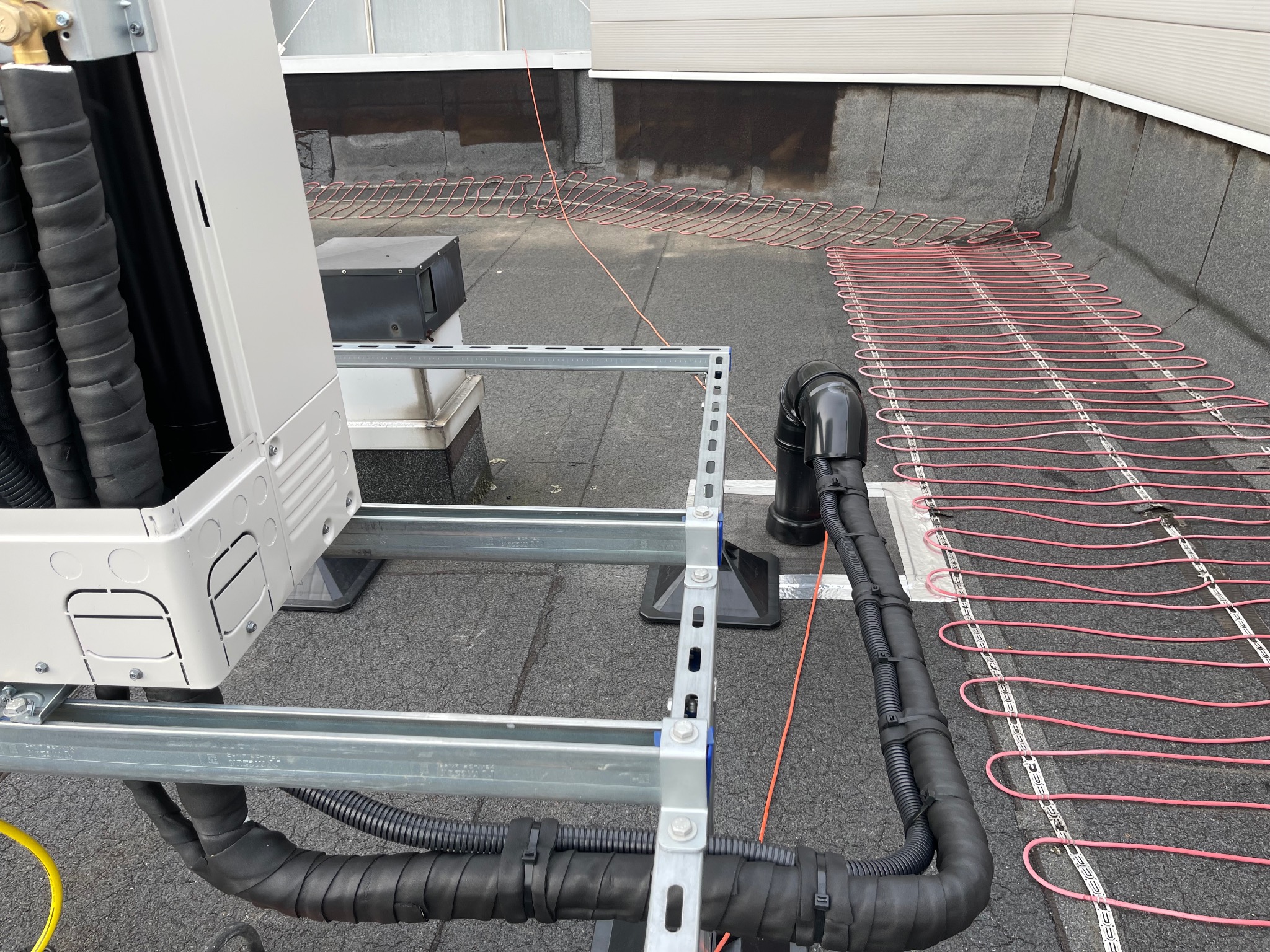 Instalacja pompy ciepła na dachu budynku z widocznymi rurami i elementami montażowymi, z bliska. Widoczne przewody grzewcze na dachu.