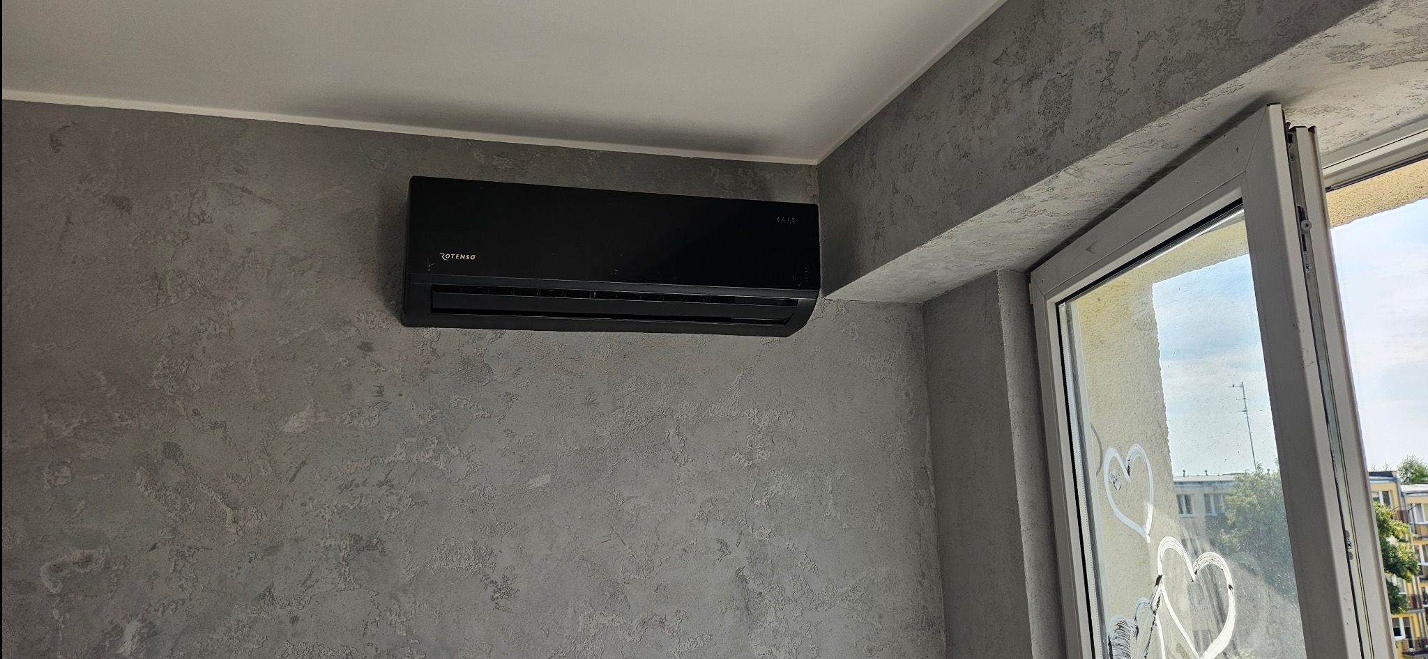 Czarny klimatyzator Rotenso zamontowany na szarej ścianie o fakturze betonu, obok otwartego okna z widokiem na zewnątrz.