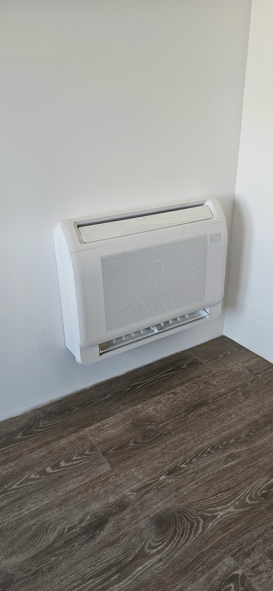 Ścienna klimatyzacja Rotenso w jasnym wnętrzu, zamontowana przy podłodze z paneli drewnopodobnych. Widok z dołu na urządzenie w rogu pomieszczenia.