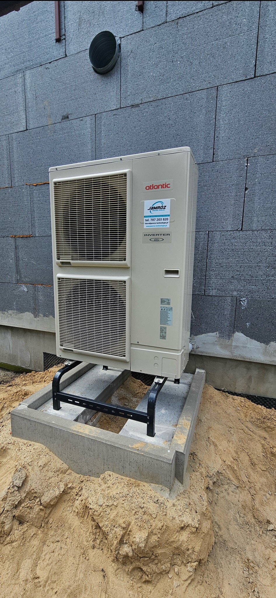 Zewnętrzna jednostka klimatyzacji Atlantic na betonowej podstawie z czarnym wspornikiem, obok ściany budynku z szarej cegły. Widoczna naklejka z danymi kontaktowymi.