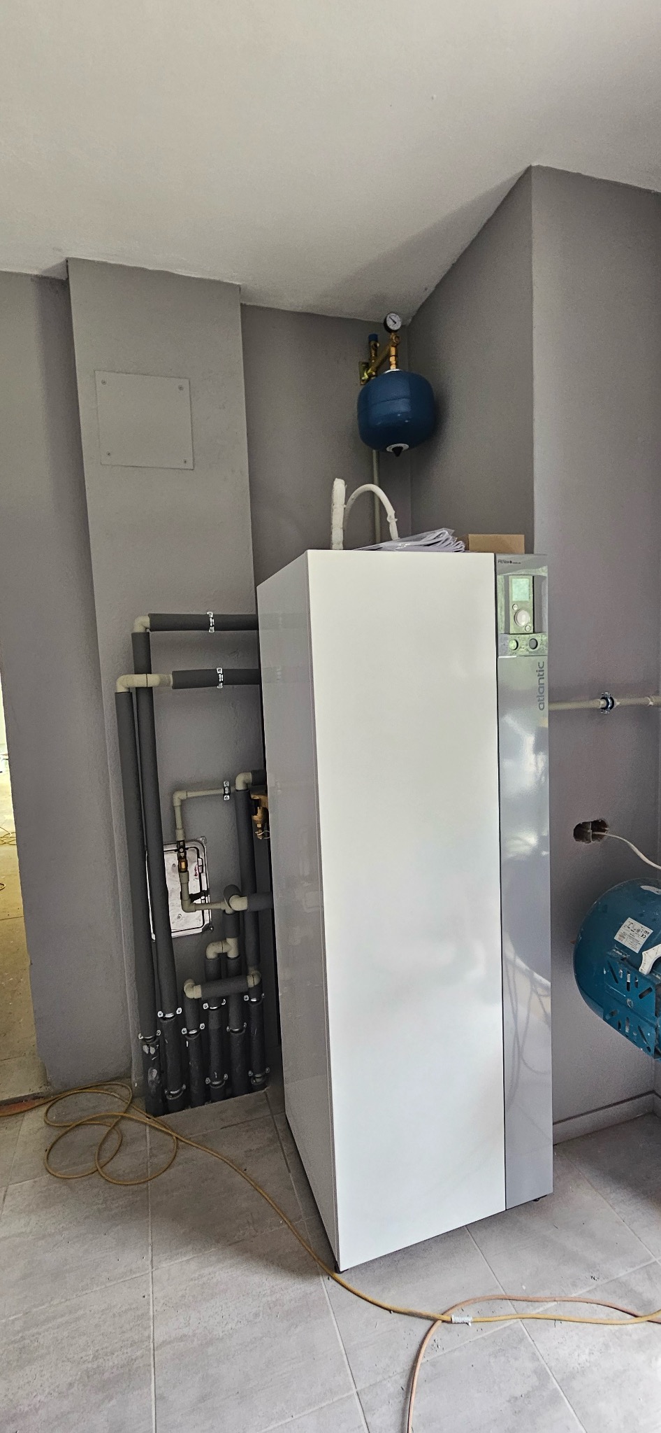 Biała pompa ciepła Atlantic w rogu szarego pomieszczenia z widoczną instalacją hydrauliczną i niebieskim zbiornikiem wyrównawczym. Widoczne przewody na podłodze.