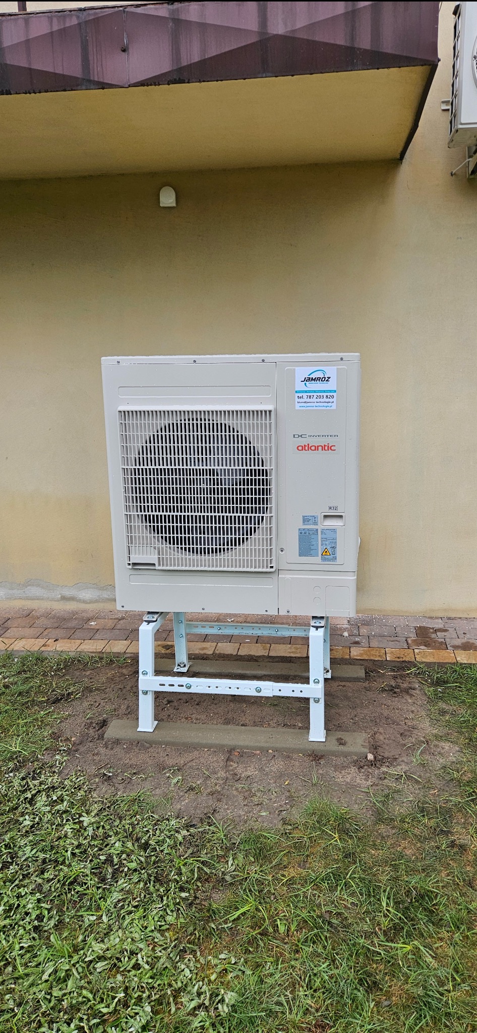 Zewnętrzna jednostka klimatyzacji Atlantic na metalowym stelażu, zamontowana przy budynku z beżową elewacją i widocznym logo firmy Jamróz. Widoczna trawa i kostka brukowa.