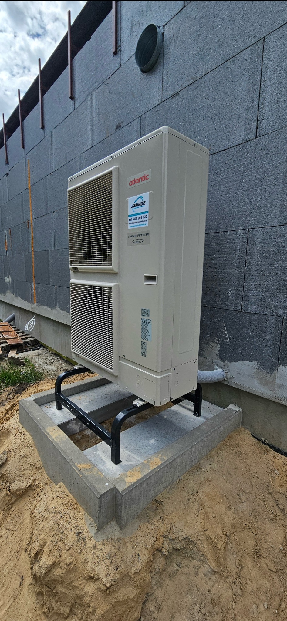 Zewnętrzna jednostka klimatyzacji Atlantic na betonowej podstawie z czarnym stelażem, przy szarym budynku z widoczną rurą wentylacyjną.