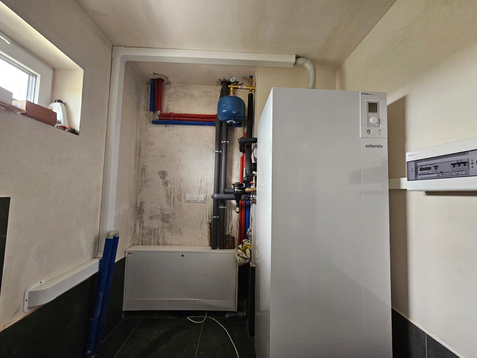 Zainstalowana pompa ciepła Atlantic w pomieszczeniu technicznym z widocznymi rurami instalacyjnymi i zbiornikiem wyrównawczym. Widoczna tablica rozdzielcza.