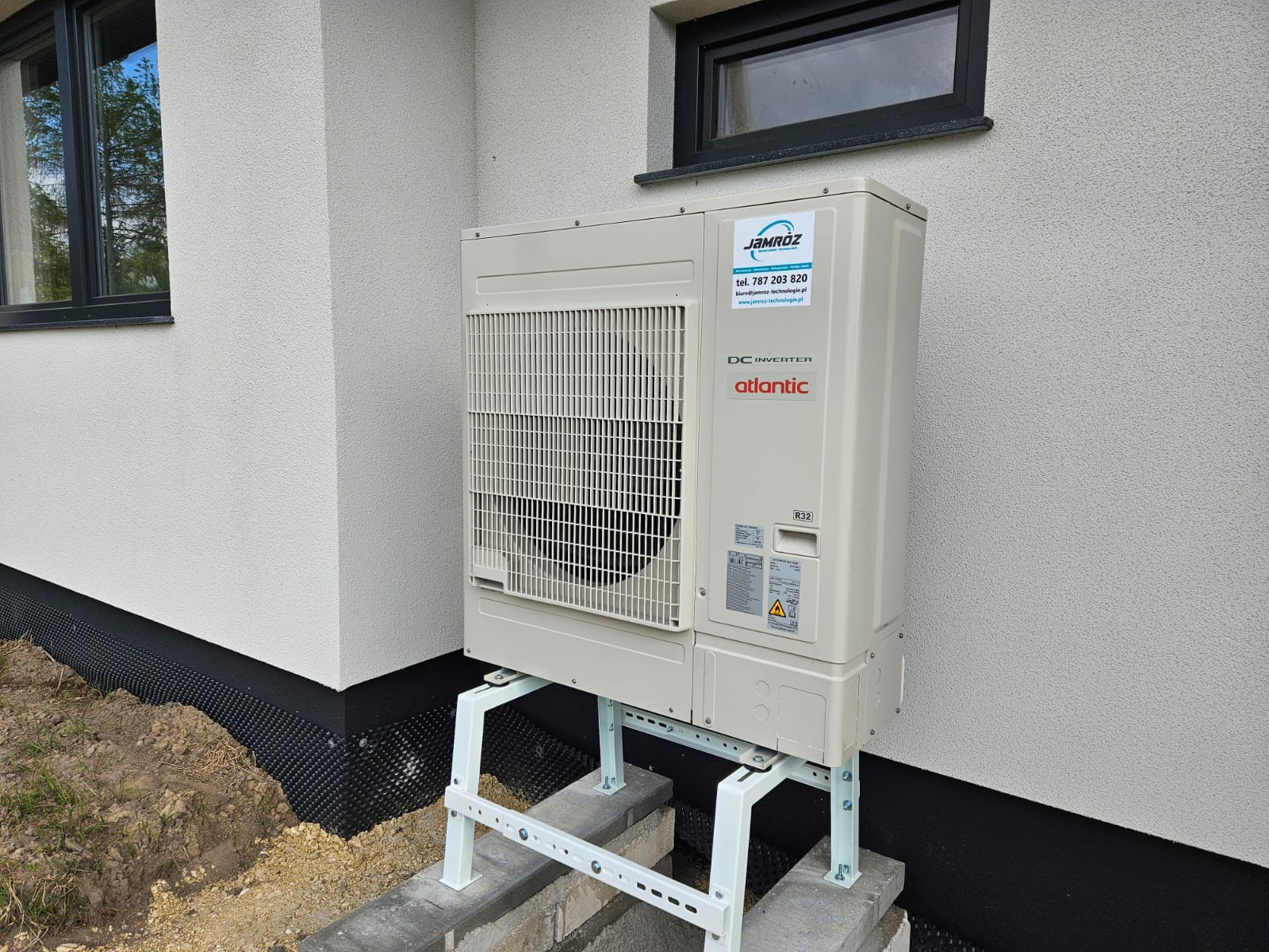 Zewnętrzna jednostka klimatyzacji Atlantic DC Inverter, zamontowana na wspornikach przy ścianie budynku z widocznym oknem i izolacją fundamentów.