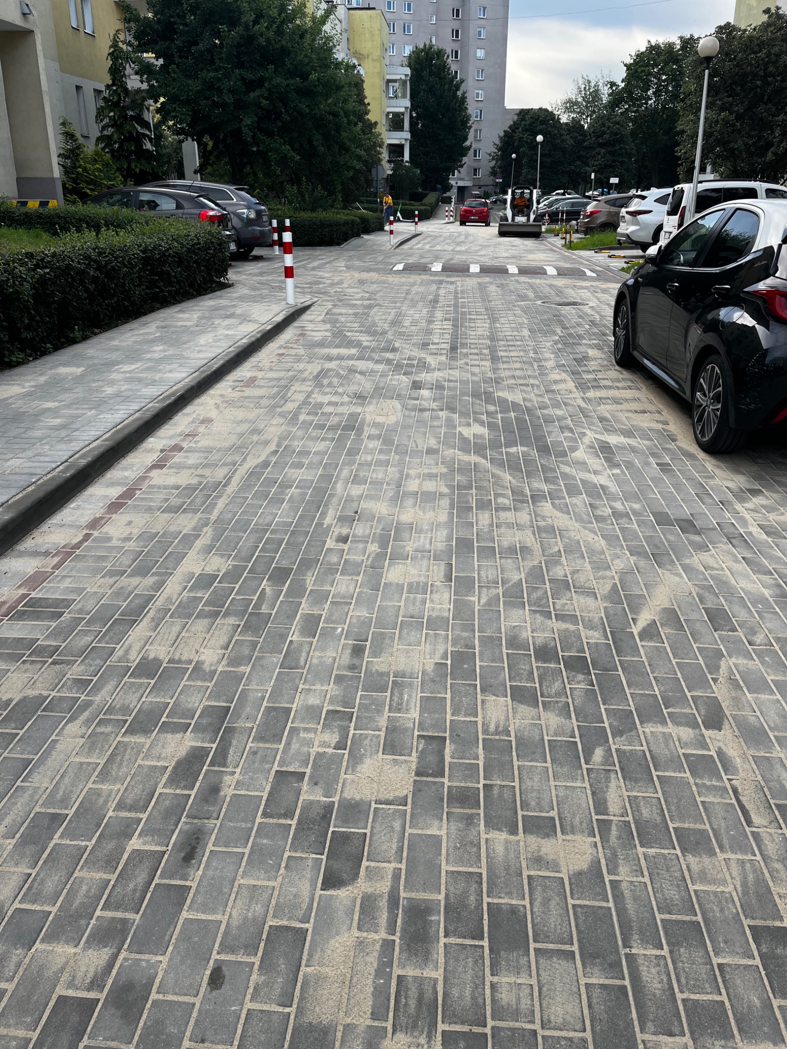 Ułożona szara kostka brukowa na drodze osiedlowej z samochodami w tle, widok z perspektywy pieszego. Słupki drogowe i progi zwalniające.