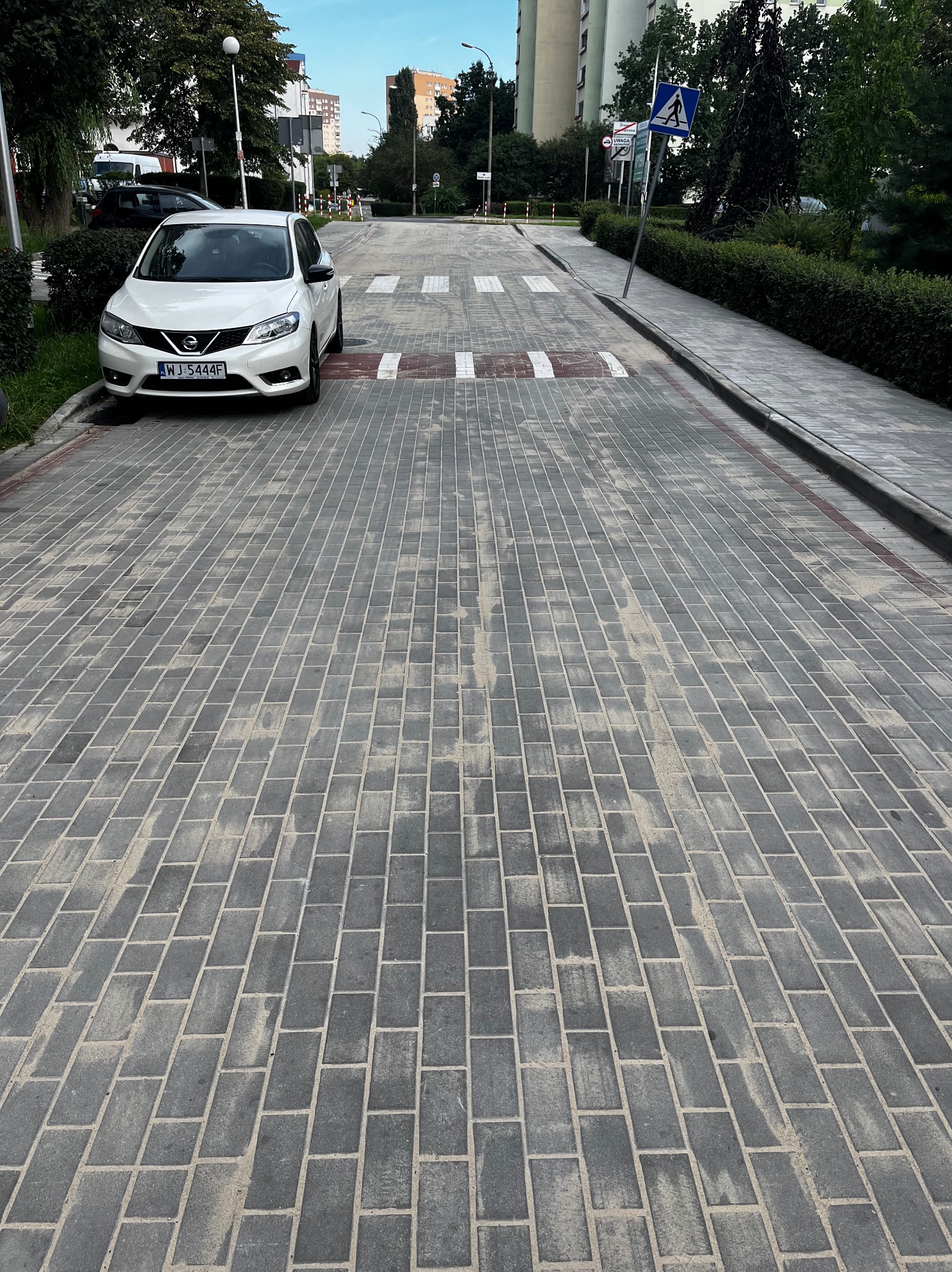 Ułożona kostka brukowa na ulicy z przejściem dla pieszych i progiem zwalniającym. Biały samochód zaparkowany obok. Widok z perspektywy pieszego.