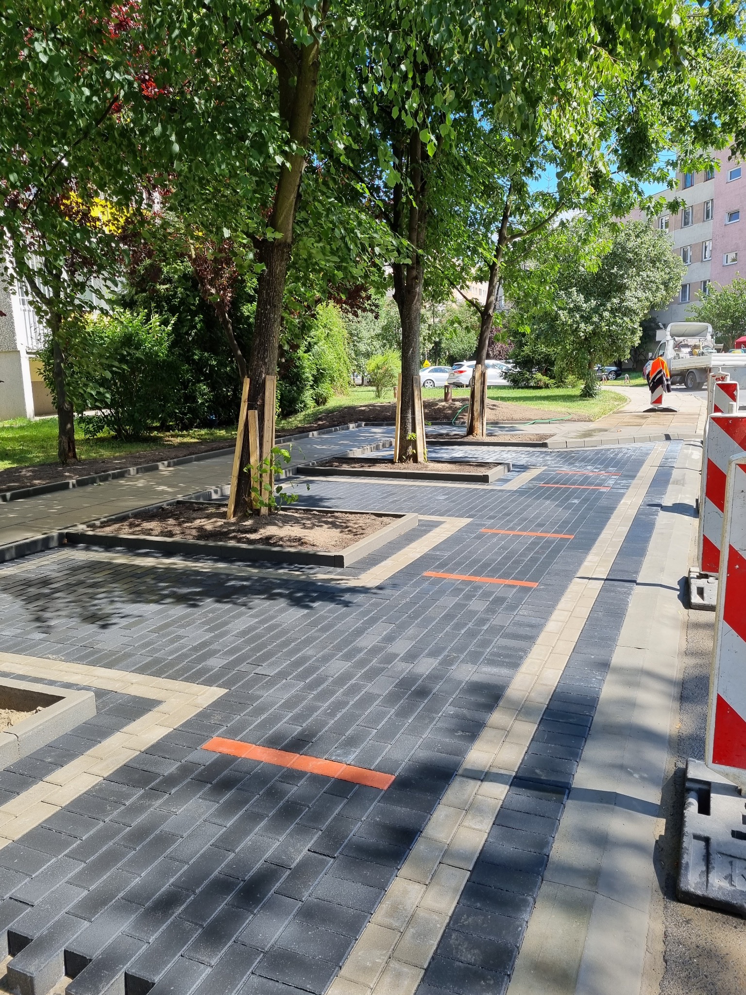 Nowa nawierzchnia z kostki brukowej w przestrzeni miejskiej, z wyznaczonymi miejscami na drzewa, otoczona zielenią i budynkami mieszkalnymi. Widoczne zabezpieczenia budowy.