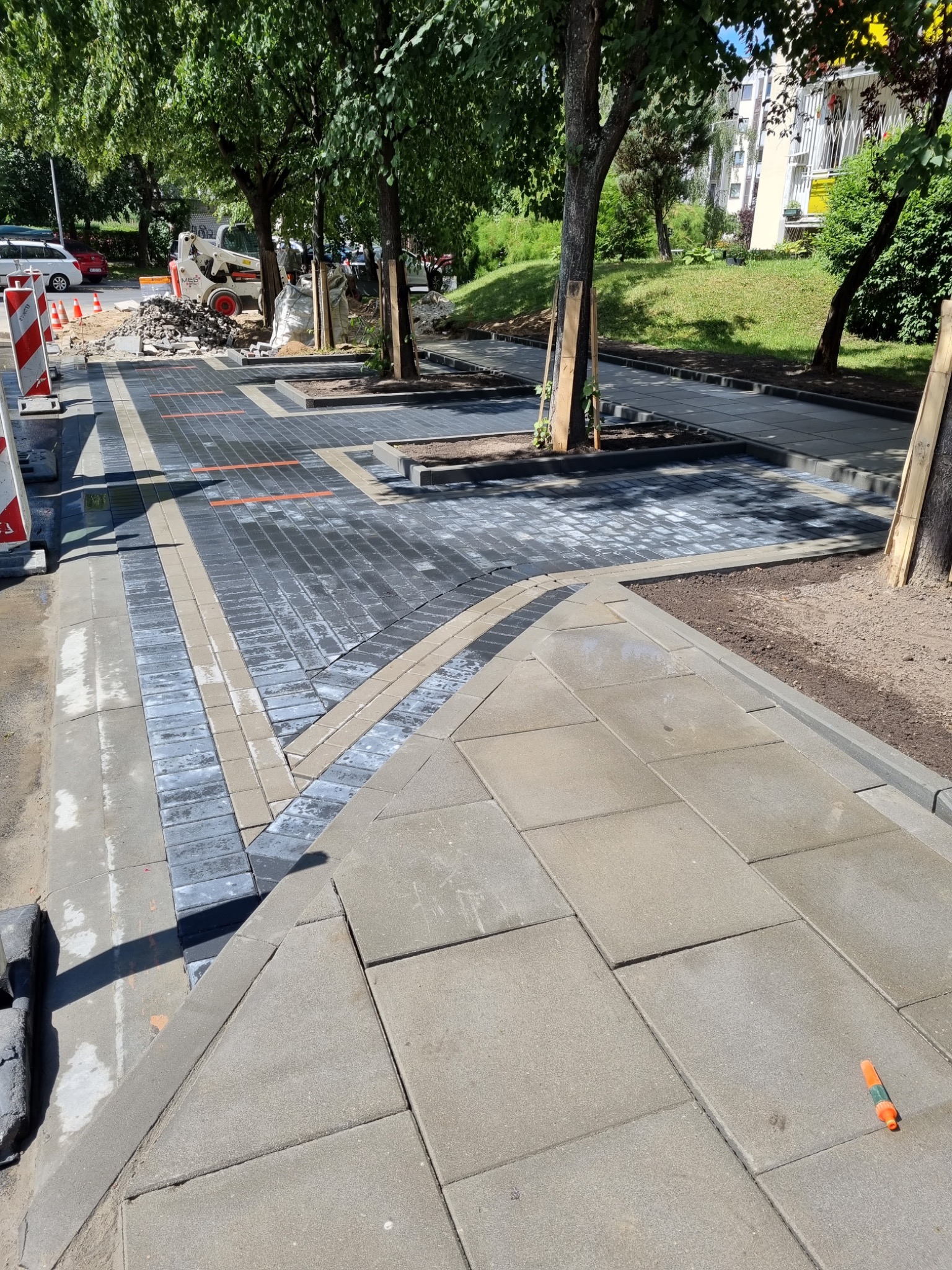 Nowa nawierzchnia z kostki brukowej w przestrzeni miejskiej, z wyznaczonymi miejscami na drzewa. Częściowo ukończona, z widocznym sprzętem budowlanym w tle.