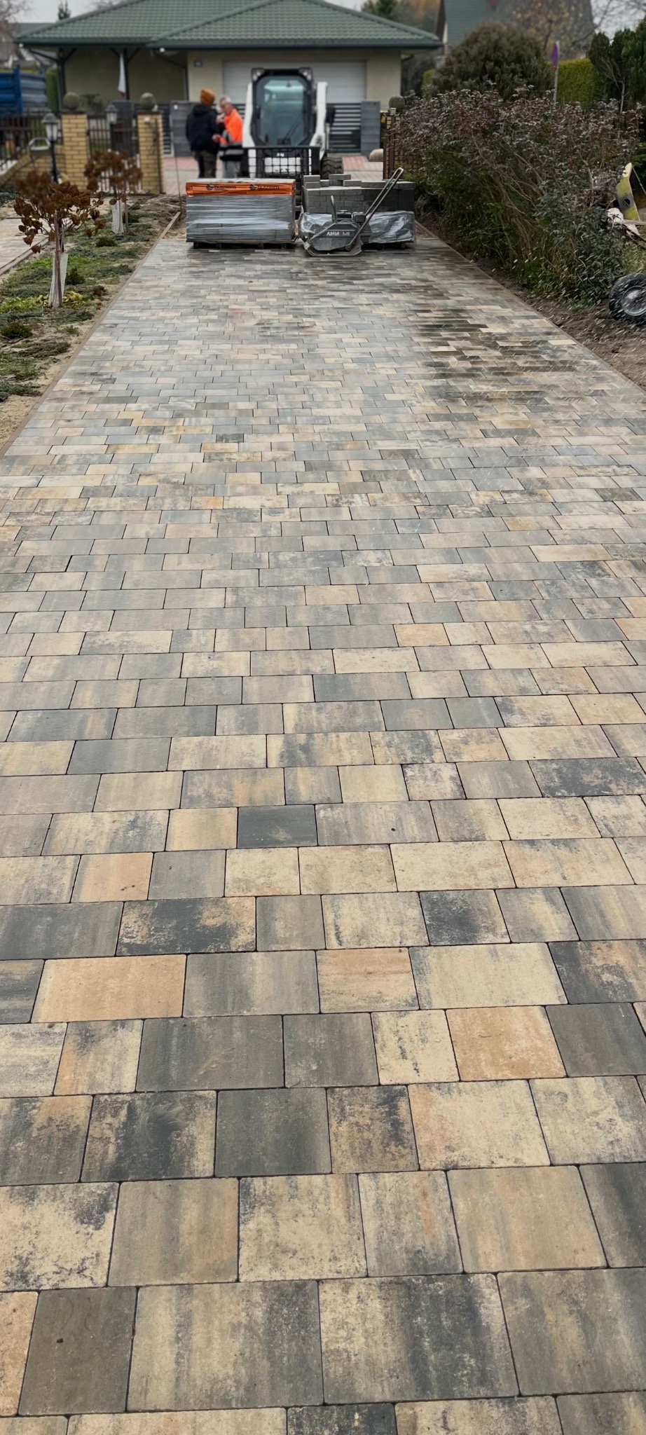 Długa, wybrukowana droga z kostki w odcieniach szarości i beżu, prowadząca do domu. W tle widoczny sprzęt budowlany i pracownicy.