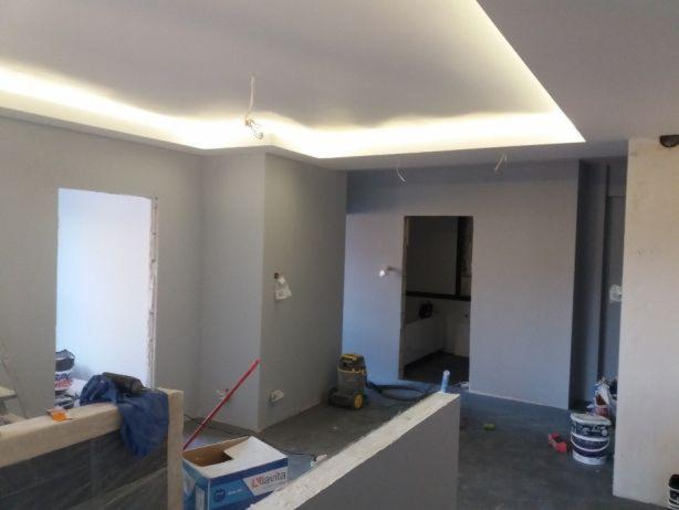 Wnętrze w trakcie remontu: szare ściany, sufit z oświetleniem LED, widoczne narzędzia i materiały budowlane, otwarty prześwit do innego pomieszczenia z białymi płytkami, odkurzacz przemysłowy w tle.