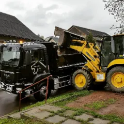 Ciężarówka MAN z grafiką lwa na kabinie, obok żółta koparko-ładowarka Komatsu WB93S podczas załadunku urobku, widoczne budynki w tle.