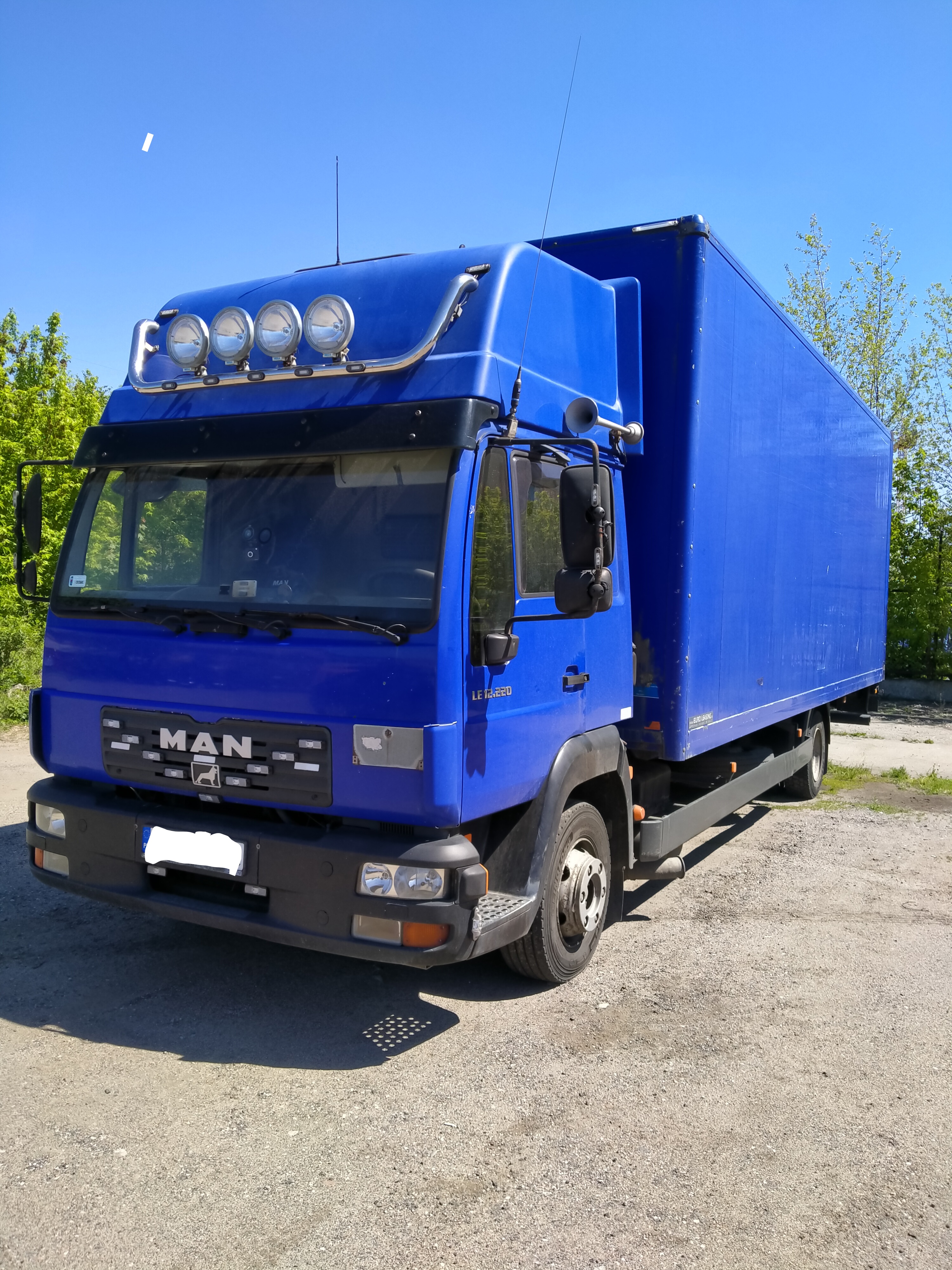 Niebieski samochód ciężarowy MAN LE 12.220 z zabudową kontenerową, wyposażony w dodatkowe reflektory na dachu, zaparkowany na szutrowym placu w słoneczny dzień.