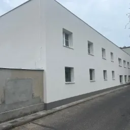 Odnowiona elewacja budynku wielorodzinnego, świeżo pomalowana na biało z szarym cokołem, widoczne okna, fragment chodnika i ulicy, ślad po naprawie na sąsiedniej ścianie.