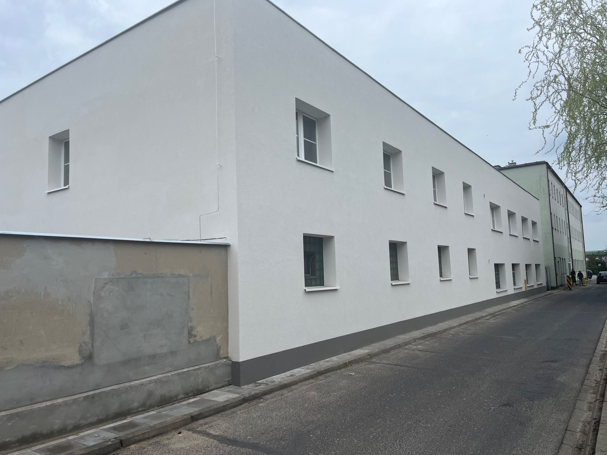 Odnowiona elewacja budynku wielorodzinnego, świeżo pomalowana na biało z szarym cokołem, widoczne okna, fragment chodnika i ulicy, ślad po naprawie na sąsiedniej ścianie.