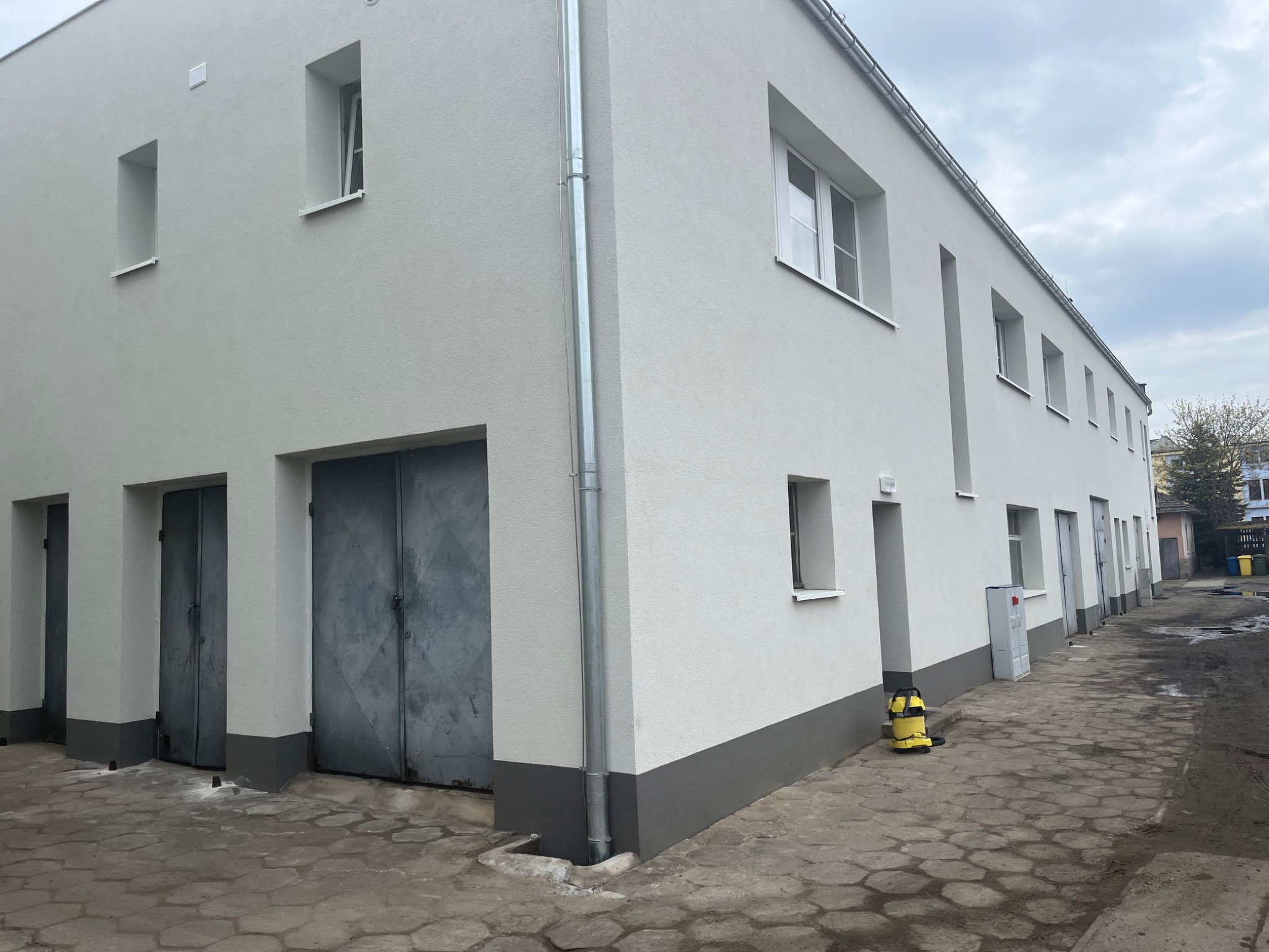 Odnowiony budynek z szarą podmurówką i nową, jasną elewacją. Widoczne metalowe drzwi garażowe, małe okna i żółty odkurzacz przemysłowy na zewnątrz.