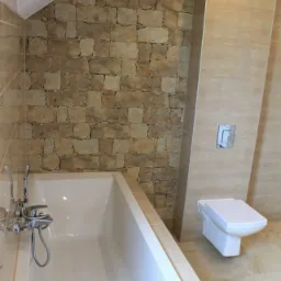 Nowoczesna łazienka z wanną, toaletą podwieszaną i ścianą wyłożoną płytkami imitującymi kamień, oświetlona punktowym światłem.