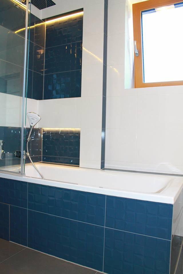 Nowoczesna łazienka z wanną obudowaną ciemnoniebieskimi, strukturalnymi płytkami, szklanym parawanem nawannowym i białymi płytkami na ścianach, oświetlona listwą LED nad wanną.