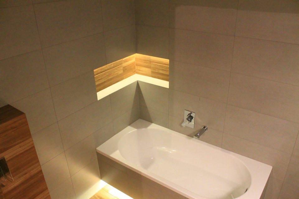 Nowoczesna łazienka z wanną obudowaną szarą płytką, wnęka z drewnianymi akcentami i podświetleniem LED oraz armaturą chromowaną.