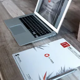 Laptop z otwartą stroną internetową firmy IT Pro, obok leży firmowy folder i długopis na drewnianym biurku, widoczny także drewniany pojemnik na długopisy.