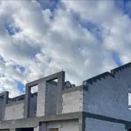 Wznoszenie ścian domu z betonu komórkowego, widoczne otwory okienne i zbrojenie pionowe, na tle błękitnego nieba z chmurami.