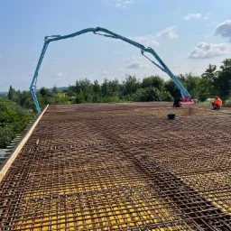 Zbrojenie stropu przygotowane do zalewania betonem, widoczna pompa do betonu z wysięgnikiem i dwóch pracowników na budowie, w tle zieleń i błękitne niebo.