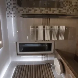 Nowoczesna łazienka z podświetlanym sufitem, grzejnikiem drabinkowym pod oknem, mozaiką nad kabiną prysznicową i umywalką wpuszczoną w blat z płytek imitujących drewno.