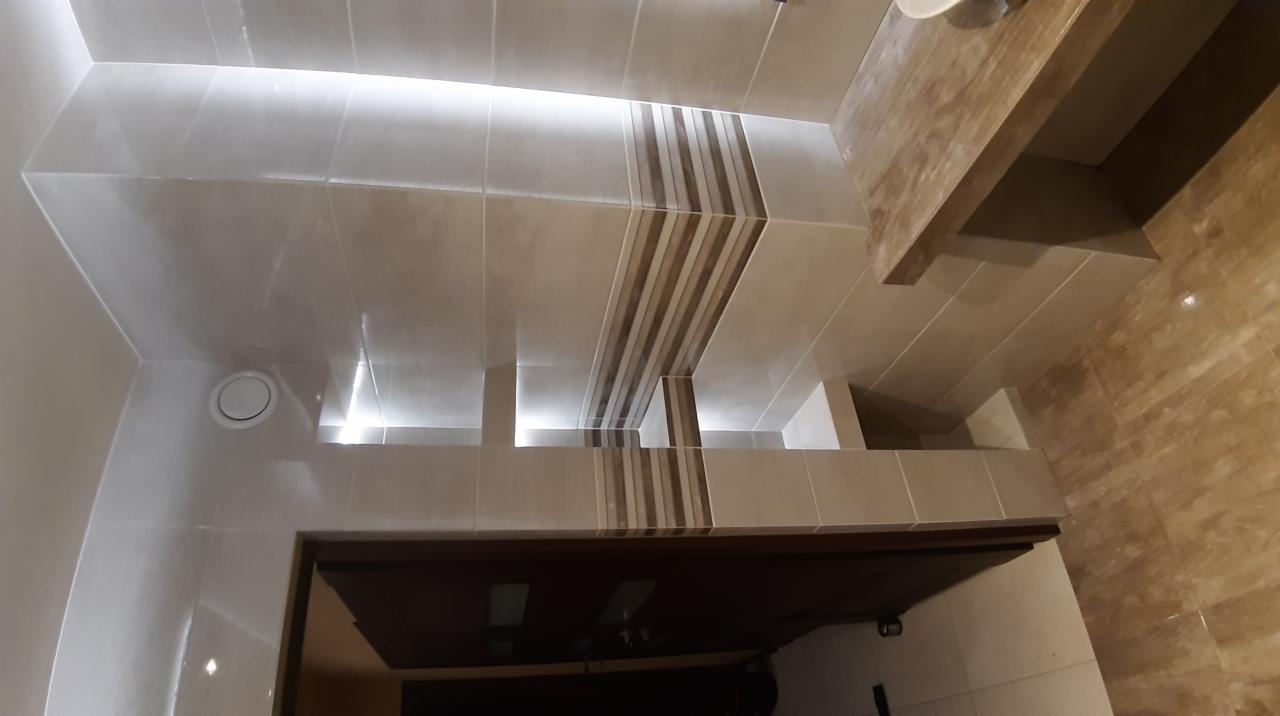 Wykończona łazienka z beżowymi płytkami imitującymi marmur na ścianach i podwieszanym sufitem z oświetleniem punktowym, widoczny fragment umywalki i lustra.