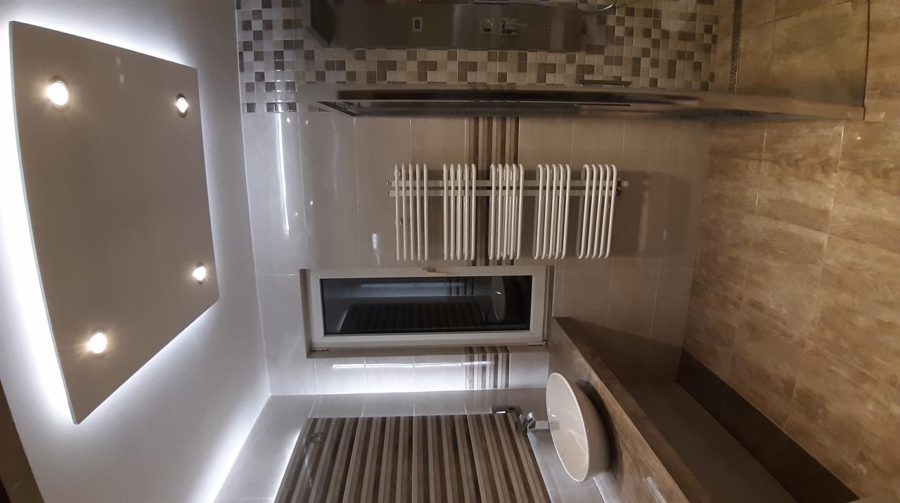 Nowoczesna łazienka z podświetlanym sufitem, grzejnikiem drabinkowym pod oknem, mozaiką nad kabiną prysznicową i umywalką wpuszczoną w blat z płytek imitujących drewno.