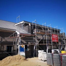 DexterBau - Wznoszony dom z ciemnej cegły pod błękitnym niebem, z widocznym rusztowaniem i materiałami budowlanymi. Etap budowy zadaszonego budynku.