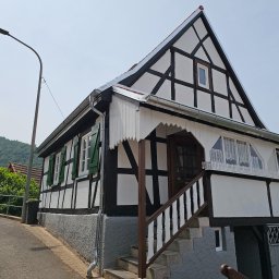 DexterBau - Dwukondygnacyjny dom w stylu szachulcowym z ciemnymi belkami i białymi ścianami, zielonymi okiennicami i schodami wejściowymi. W tle zalesione wzgórza.