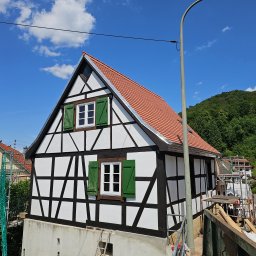 DexterBau - Dom w stylu szachulcowym z czerwoną dachówką i zielonymi okiennicami, na tle błękitnego nieba z chmurami i zielonego wzgórza. Widoczne elementy rusztowania.