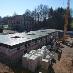 DexterBau - Budowa domu z keramzytu: ściany z pustaków, strop, rusztowania, żuraw. W tle budynek, drzewa i błękitne niebo. Widok z góry na plac budowy w słoneczny dzień.