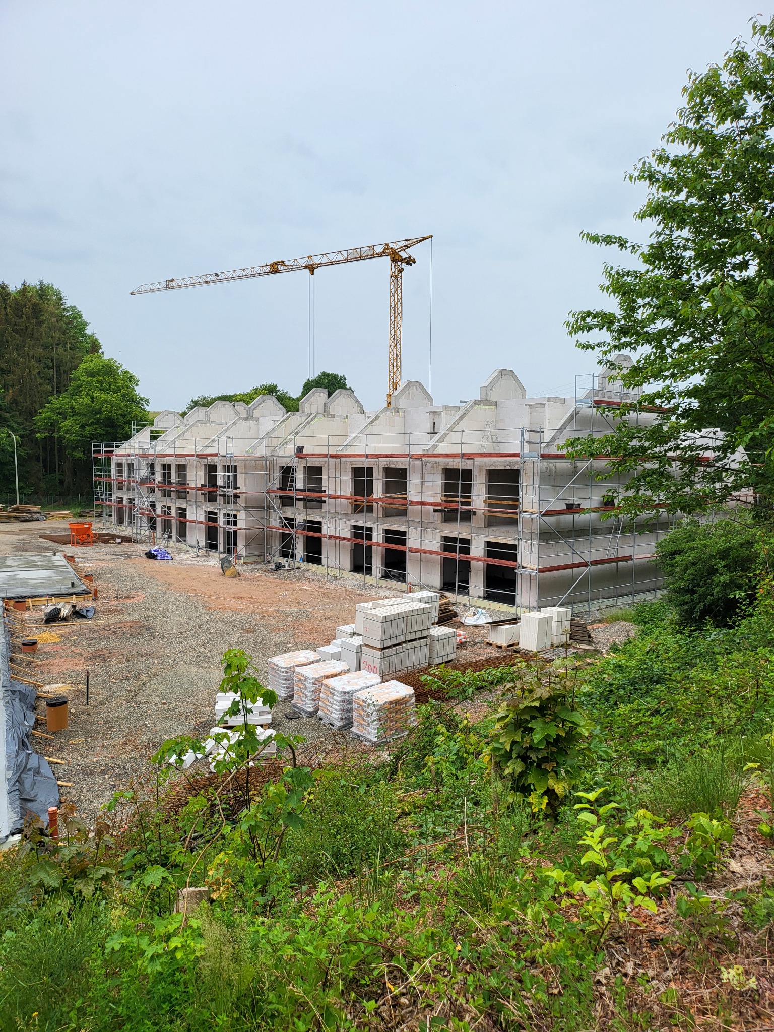 Wznoszenie dwukondygnacyjnego domu z białych bloczków, widoczne rusztowania i żuraw budowlany. Na pierwszym planie składowane materiały budowlane i zieleń.