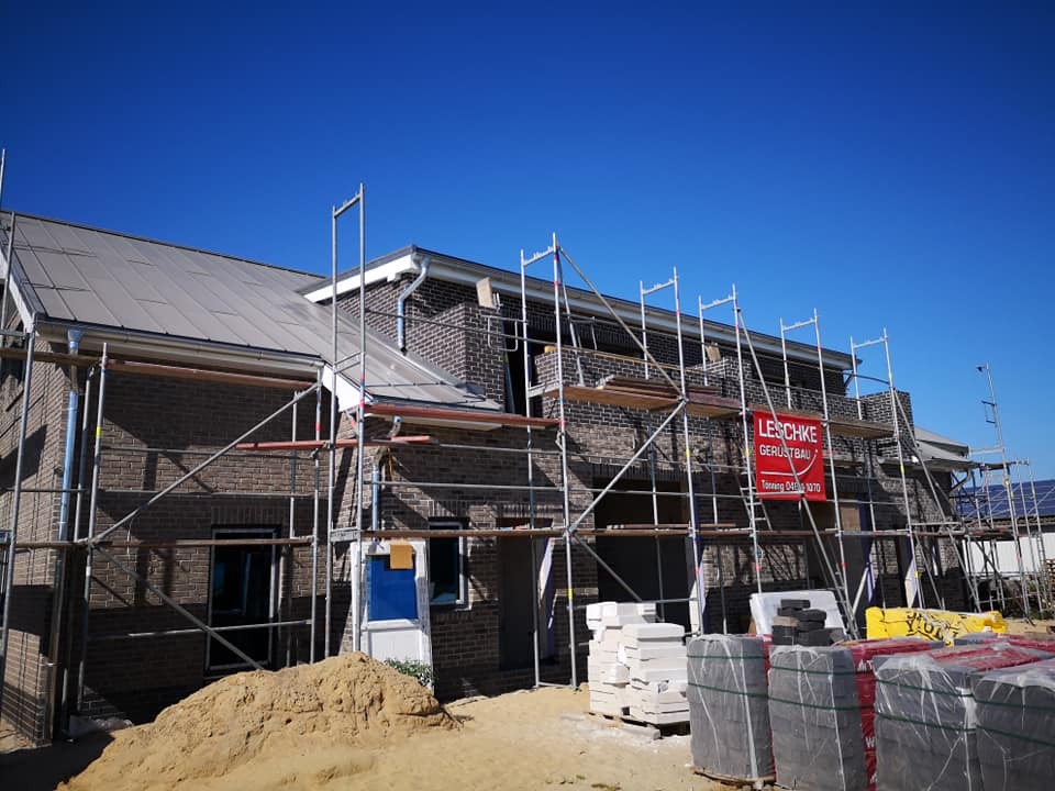 Wznoszony dom z ciemnej cegły pod błękitnym niebem, z widocznym rusztowaniem i materiałami budowlanymi. Etap budowy zadaszonego budynku.