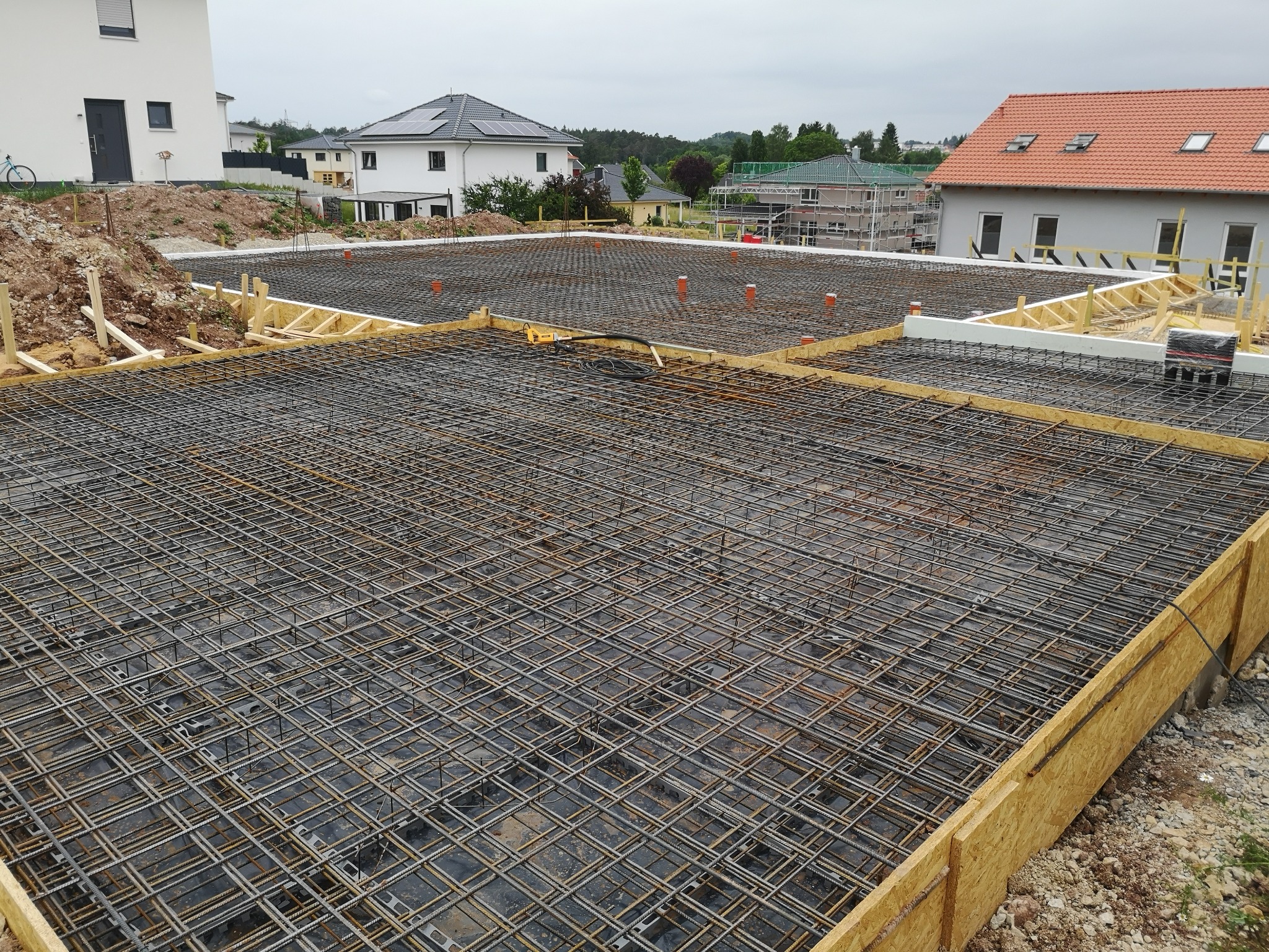 Zbrojenie fundamentów domu jednorodzinnego z widocznymi elementami szalunku i instalacji. W tle zabudowa mieszkalna i pochmurne niebo.