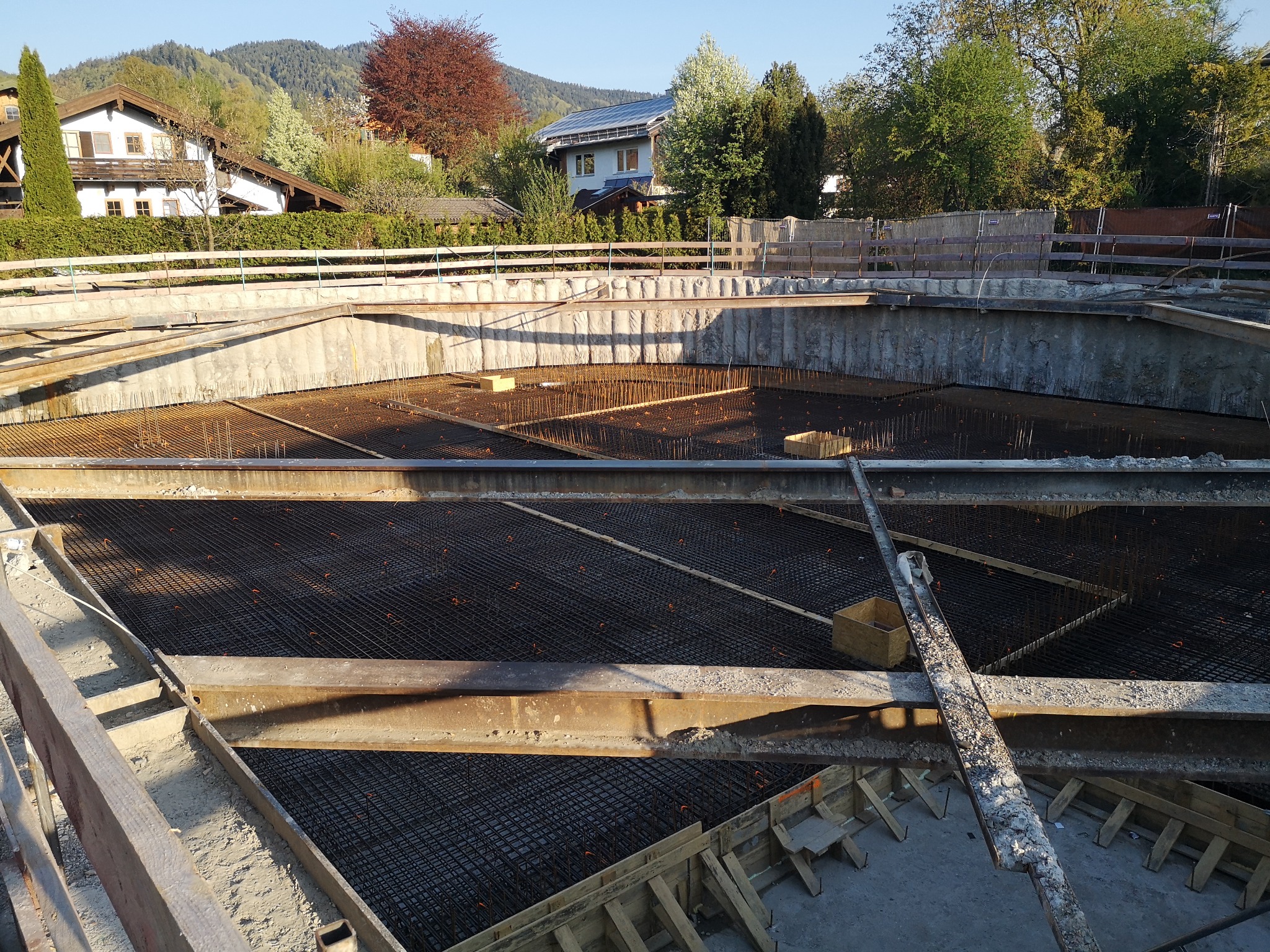 Zbrojenie fundamentów domu jednorodzinnego z widoczną siatką stalową, szalunkiem i elementami konstrukcyjnymi. W tle zabudowa mieszkalna i zieleń.