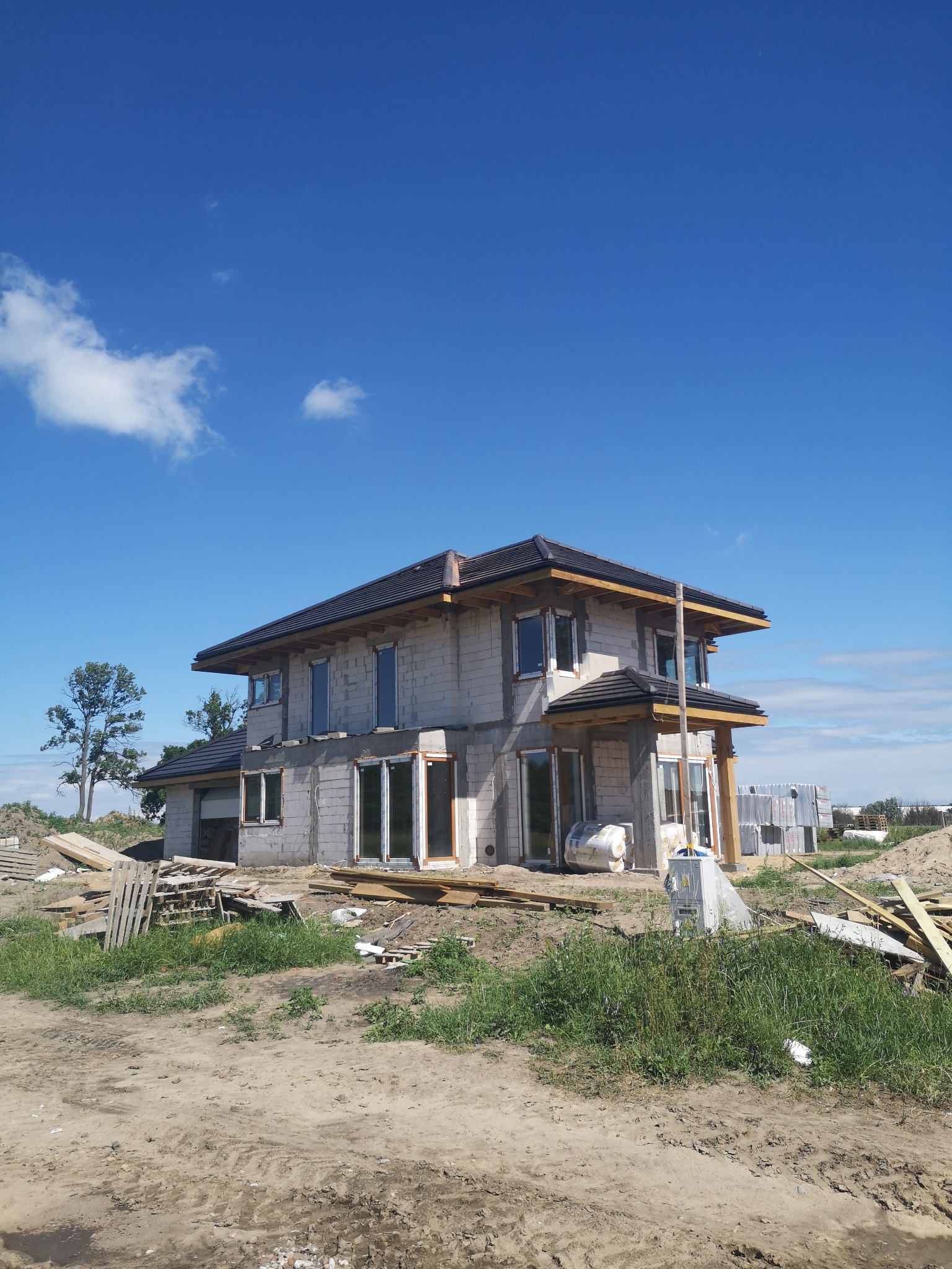 Dwukondygnacyjny dom w stanie surowym otwartym z ciemną dachówką, widoczne materiały budowlane i niezagospodarowany teren wokół. Błękitne niebo z pojedynczymi chmurami.