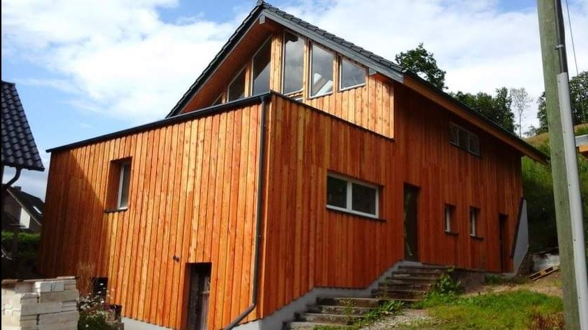 Nowoczesny dom szkieletowy z drewnianą elewacją w odcieniach brązu, z dużymi oknami na poddaszu i schodami wejściowymi. Widok z zewnątrz w słoneczny dzień.