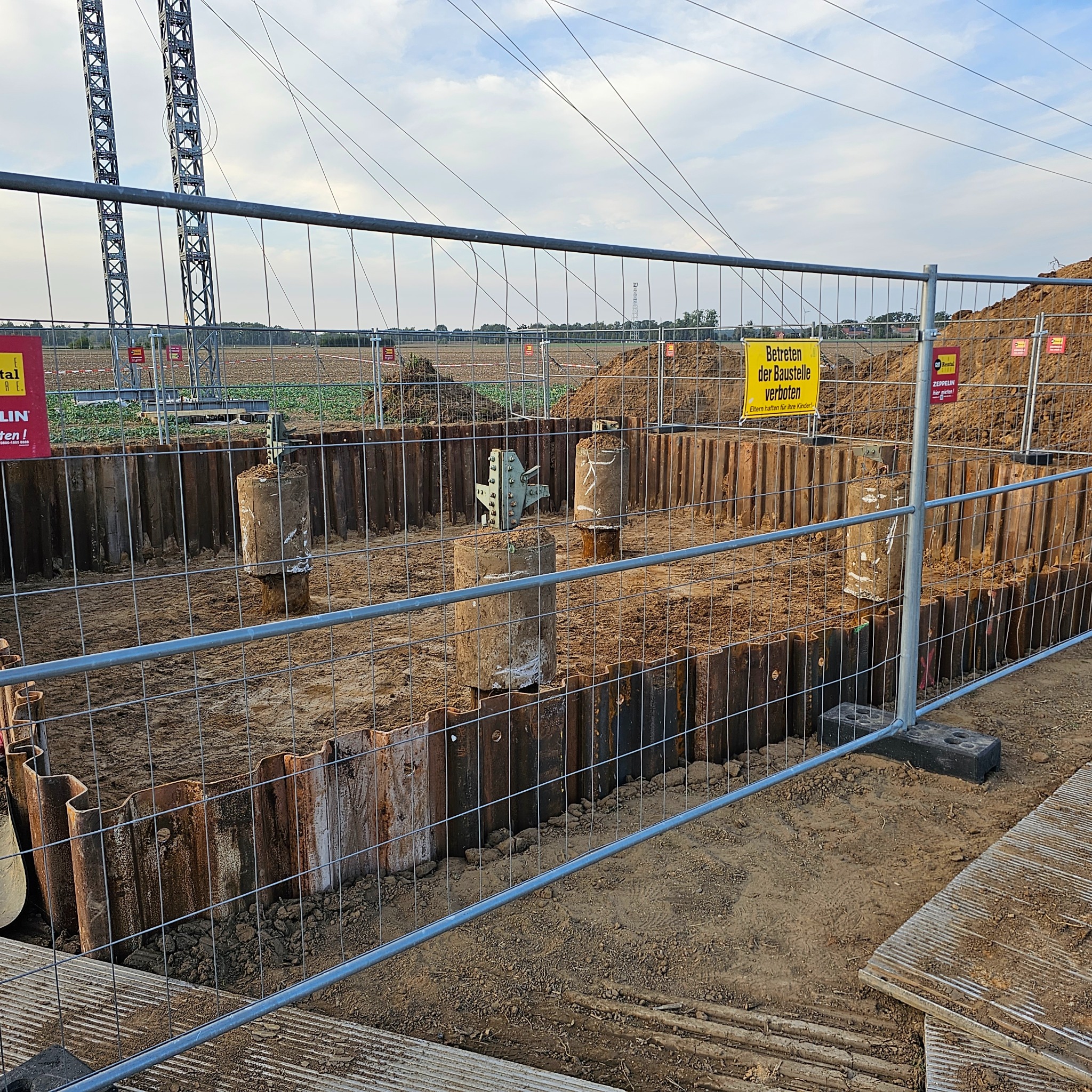 Teren budowy z zabezpieczeniami wykopu, widoczne stalowe ścianki Larsena i betonowe pale. Na pierwszym planie siatka ogrodzeniowa i tablice informacyjne.