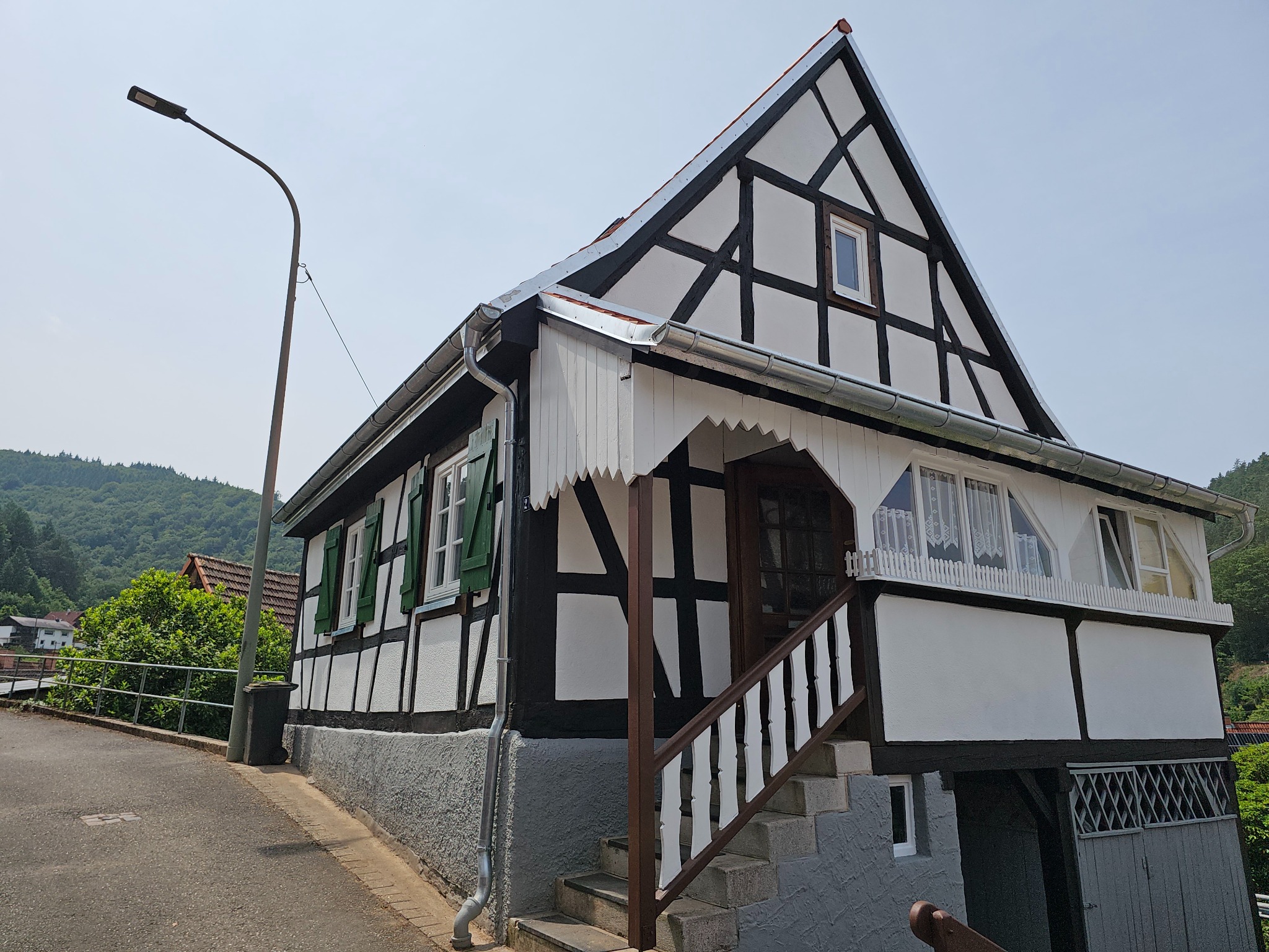 Dwukondygnacyjny dom w stylu szachulcowym z ciemnymi belkami i białymi ścianami, zielonymi okiennicami i schodami wejściowymi. W tle zalesione wzgórza.