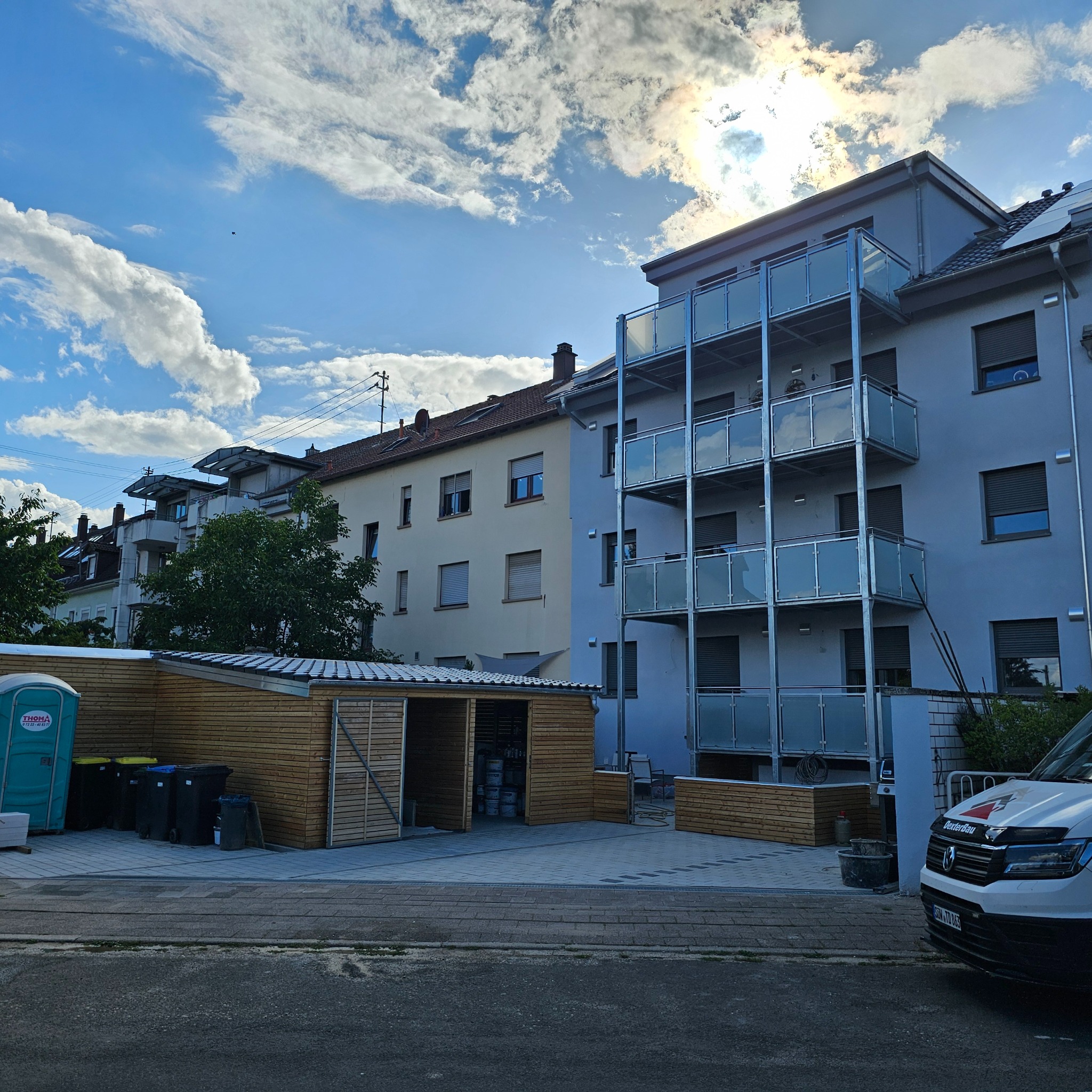 Nowoczesny budynek wielorodzinny z balkonami o szklanych balustradach, widoczny z poziomu ulicy. Na pierwszym planie drewniana wiata i zaparkowany samochód dostawczy.