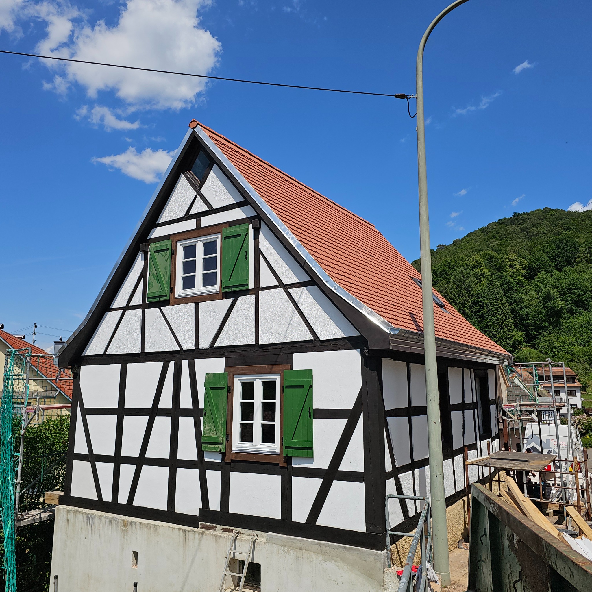 Dom w stylu szachulcowym z czerwoną dachówką i zielonymi okiennicami, na tle błękitnego nieba z chmurami i zielonego wzgórza. Widoczne elementy rusztowania.