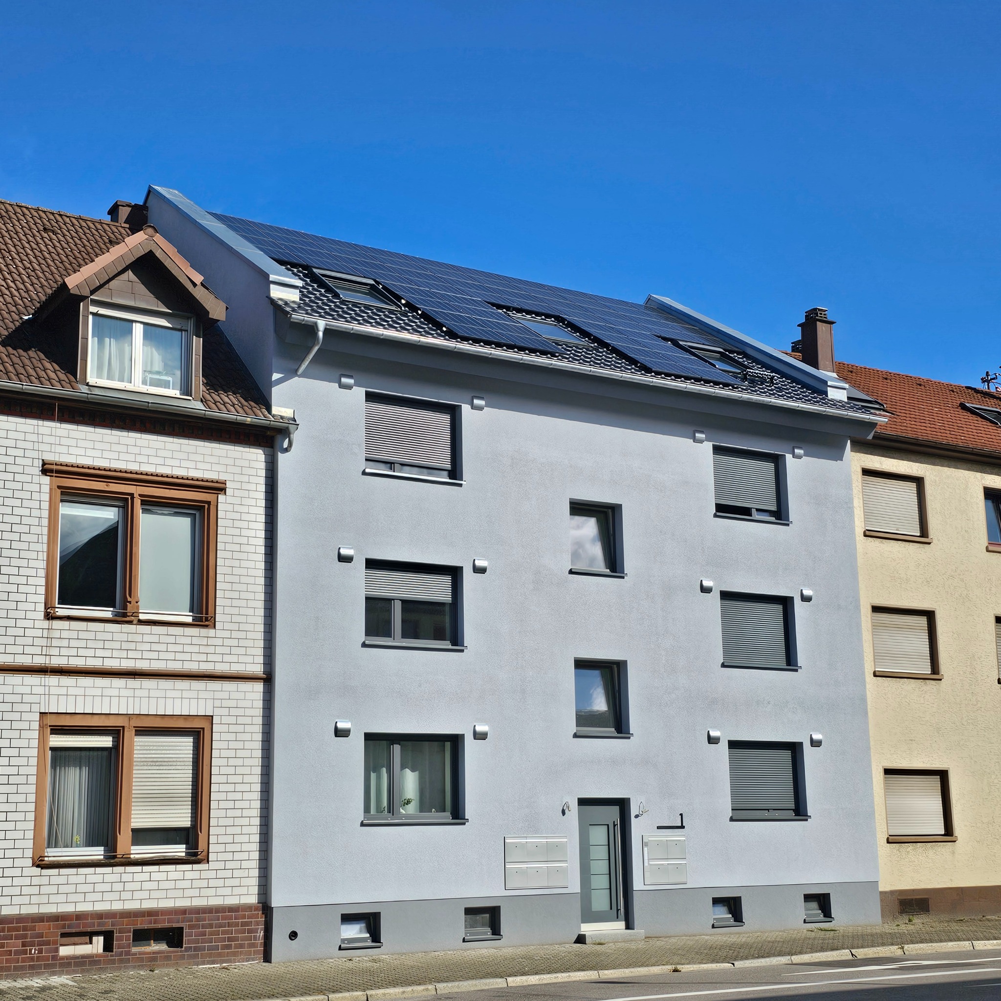 Szary, trzypiętrowy budynek z panelami słonecznymi na dachu, sąsiadujący z budynkami o różnej architekturze i kolorystyce, widziany w słoneczny dzień.