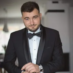 Elegancki mężczyzna w czarnym smokingu i muszce, z zegarkiem na nadgarstku, spogląda w obiektyw z lekkim uśmiechem. Jasne tło z elementami wystroju wnętrza.