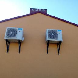 Coldmark - Dwa identyczne klimatyzatory Midea zamontowane na czarnych wspornikach na pomarańczowej ścianie budynku z ciemnoczerwonym dachem i kominem na tle błękitnego nieba.