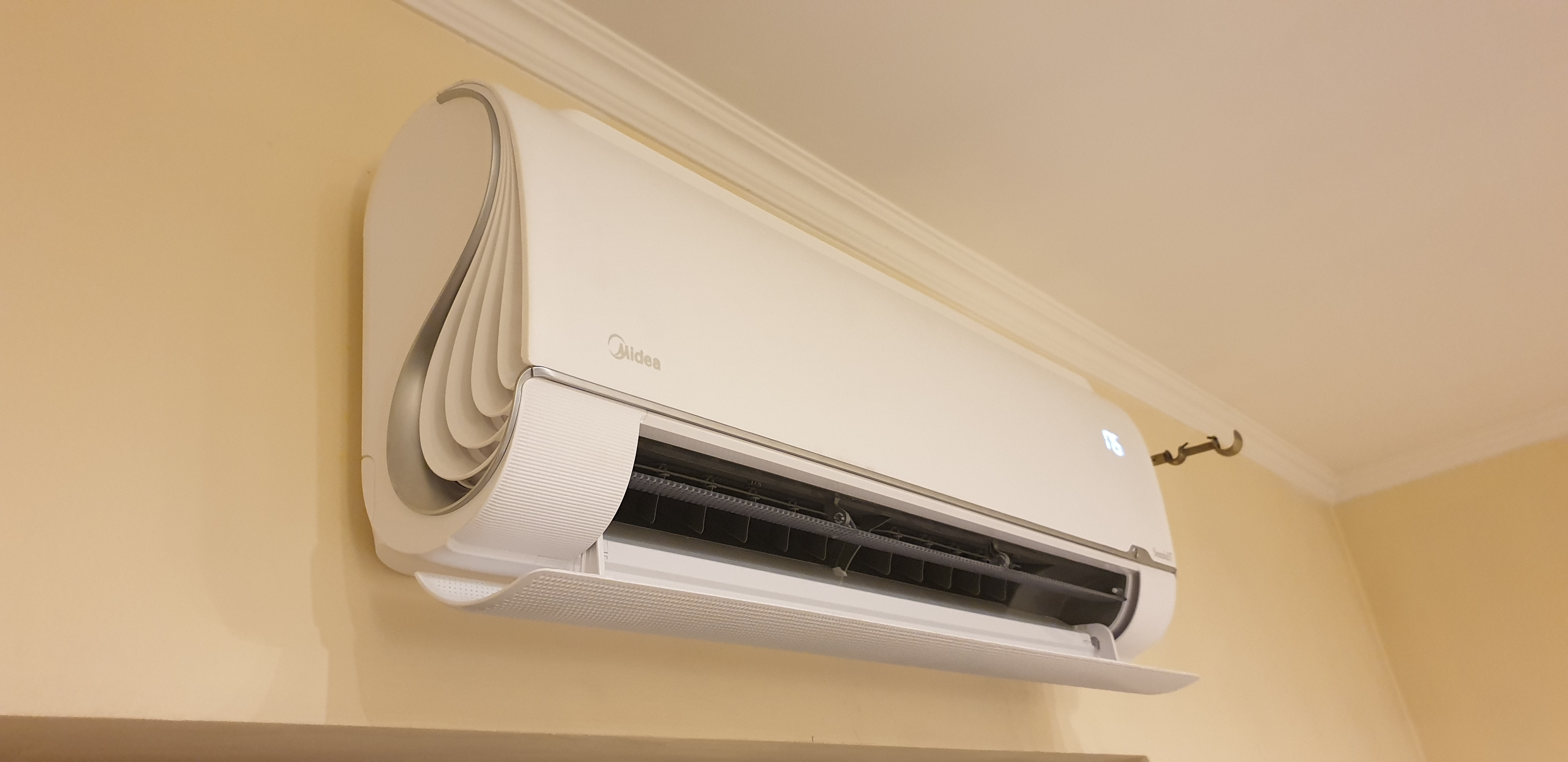 Nowoczesny, biały klimatyzator Midea zamontowany na ścianie w pomieszczeniu. Widoczny panel sterowania z wyświetlaną temperaturą. Dekoracyjne wykończenie.